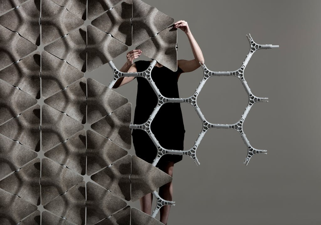 Bilden visar ett modernt modulärt akustiskt avdelarsystem med ett texturerat grått filtliknande material arrangerat i ett geometriskt mönster. Strukturen har triangulära utsprång och visas med en hexagonal ramkomponent. En person är delvis synlig och interagerar med installationen, vilket antyder dess skala och användning som rumsavdelare snarare än väggmonterade paneler.